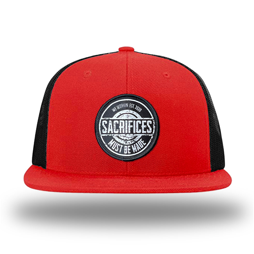 Red/Black WeWorkin hat—Richardson 511 brand snapback, flatbill trucker hat style. WeWorkin "SACRIFICES Must Be Made" circular woven patch, with black/white thread colors and black merrowed edge, is centered on the front panels.