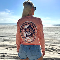 Woman pictured at the ocean, from the back, in a WE WORKIN ultralight weight, coral color long-sleeved tee—Full back is screenprinted with "HOOKED ON HARD WORK | WE WORKIN | MMXX" text encircling a skeletal Marlin fish graphic, printed in navy and white ink.