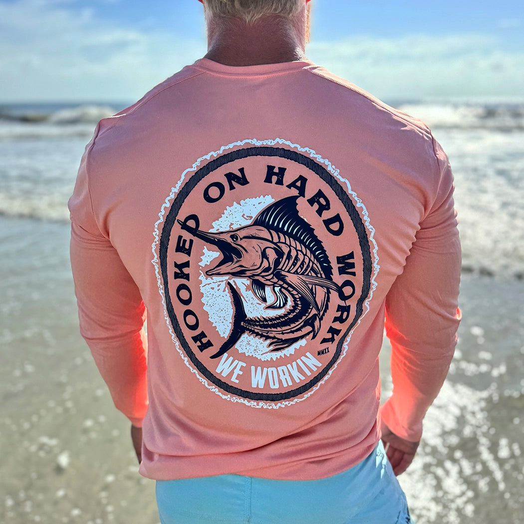 Man pictured at the ocean, from the back, in a WE WORKIN ultralight weight, coral color long-sleeved tee—Full back is screenprinted with "HOOKED ON HARD WORK | WE WORKIN | MMXX" text encircling a skeletal Marlin fish graphic, printed in navy and white ink.