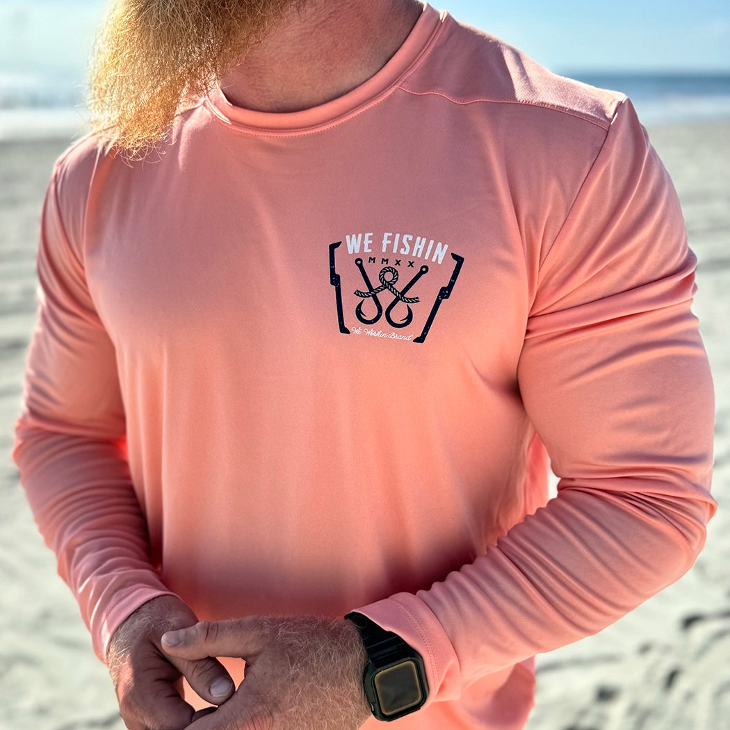 Man pictured outdoors, from the front, in a WE WORKIN ultralight weight, coral color long-sleeved tee—left chest "pocket area" screenprinted with "WE FISHIN | MMXX | We Workin Brand" text and 2 fish hooks/line graphic, printed in navy and white ink.