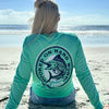 Woman pictured at the ocean, from the back, in a WE WORKIN ultralight weight, seafoam green color long-sleeved tee—Full back is screenprinted with "HOOKED ON HARD WORK | WE WORKIN | MMXX" text encircling a skeletal Marlin fish graphic, printed in navy and white ink.