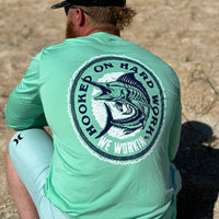 Man pictured at the ocean, from the back, in a WE WORKIN ultralight weight, seafoam green color long-sleeved tee—Full back is screenprinted with "HOOKED ON HARD WORK | WE WORKIN | MMXX" text encircling a skeletal Marlin fish graphic, printed in navy and white ink.