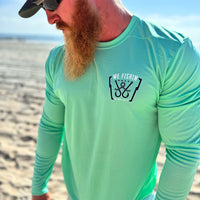 Man pictured outdoors, from the front, in a WE WORKIN ultralight weight, seafoam green color long-sleeved tee—left chest "pocket area" screenprinted with "WE FISHIN | MMXX | We Workin Brand" text and 2 fish hooks/line graphic, printed in navy and white ink.