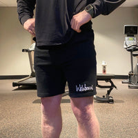 Man standing in gym, pictured from front with the We Workin men's shorts in BLACK. "WEWORKIN" script logo is embroidered in white thread on the left leg, bottom hem, on front. [Graphic approx 4" wide.]
