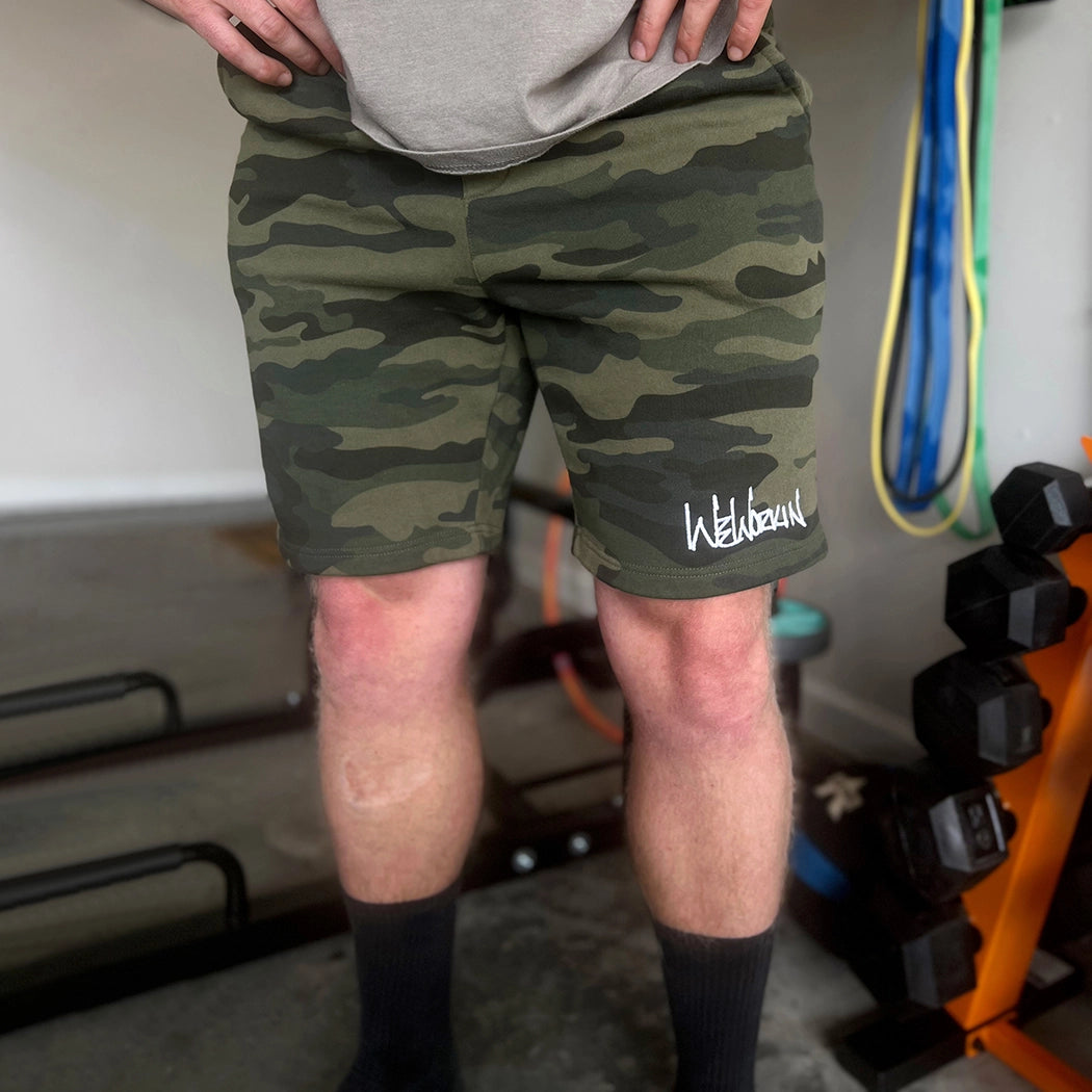 Man standing in gym, pictured from front with the We Workin men's shorts in FOREST CAMO. "WEWORKIN" script logo is embroidered in white thread on the left leg, bottom hem, on front. [Graphic approx 4" wide.]