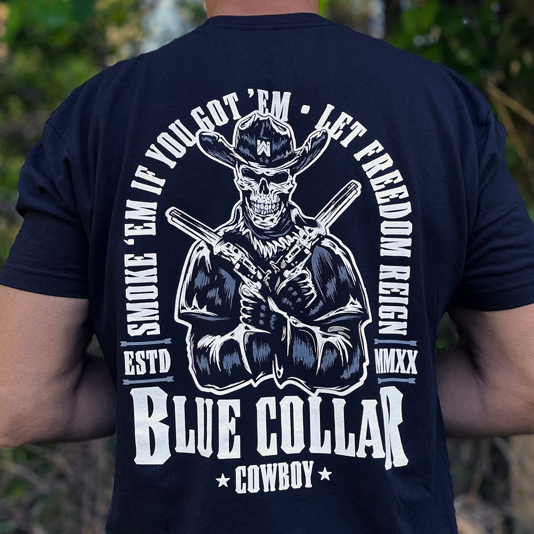 Man wearing a WW black tee, shown from back. "SMOKE 'EM IF YOU GOT 'EM • LET FREEDOM REIGN" text arching around the top and sides of a skeleton cowboy in a duster as center image. BLUE COLLAR COWBOY is along the bottom of the graphic, printed large on the center/upper back. Printed in grey and white ink.