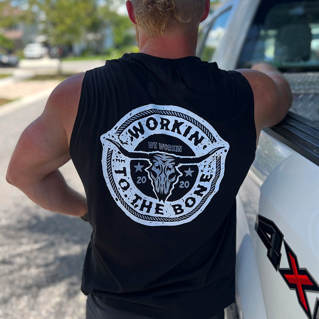 Man pictured from back, wearing a We Workin muscle tank in black. "WORKIN TO THE BONE" text circular graphic with a cow skull in the center is printed large in the center/upper back, in grey and white ink.
