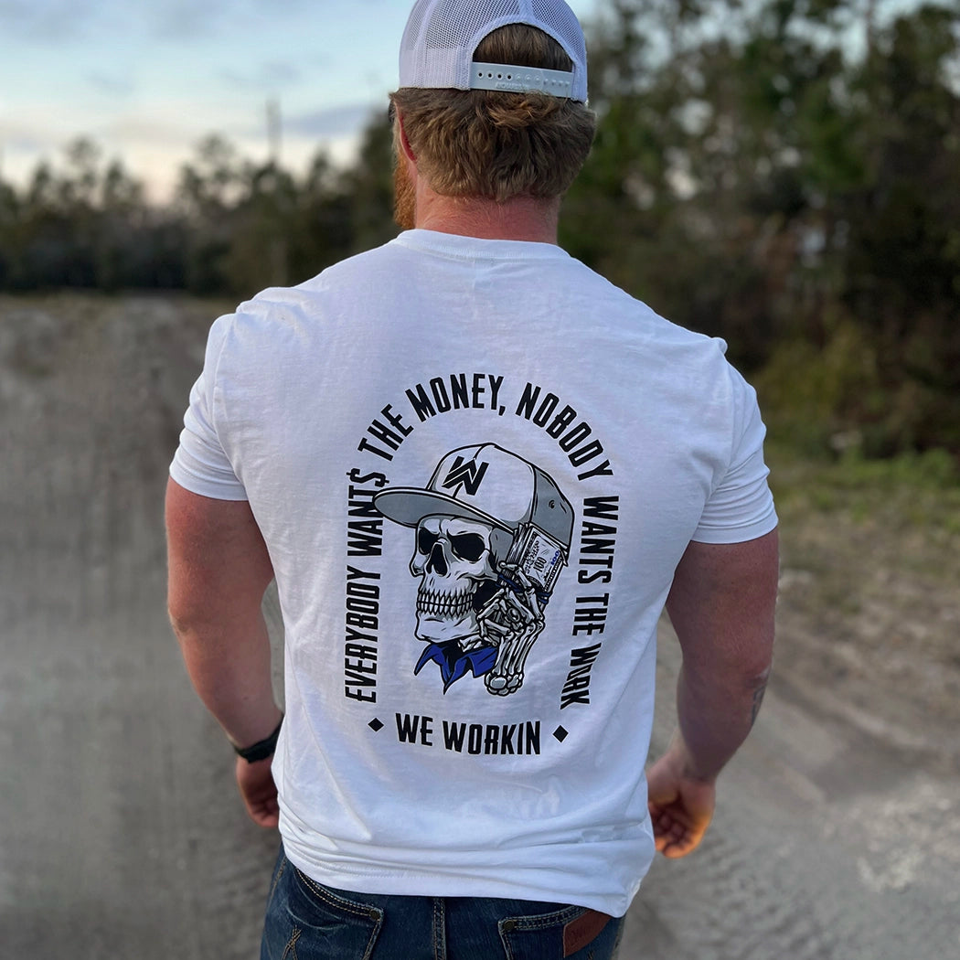 Man pictured from back wearing a WE WORKIN White tee with the large graphic "EVERYBODY WANT$ THE MONEY, NOBODY WANTS THE WORK" tagline and WE WORKIN text, surrounding a Skull wearing a hat and holding a money bundle to his ear on back. Skull graphic in color, text in black.