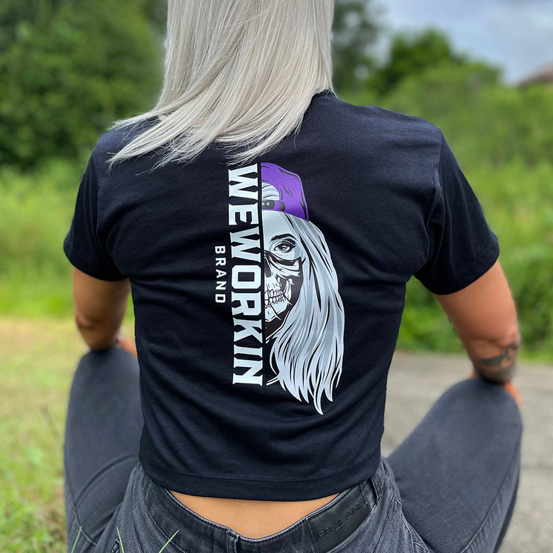 Woman wearing a We Workin Women's short-sleeve cropped tee in black—with a large imprint of our "WEWORKIN BRAND vertical text and Half Skull Woman with Hat" design in Black/White/Grey (hat graphic highlighted in Purple ink.)