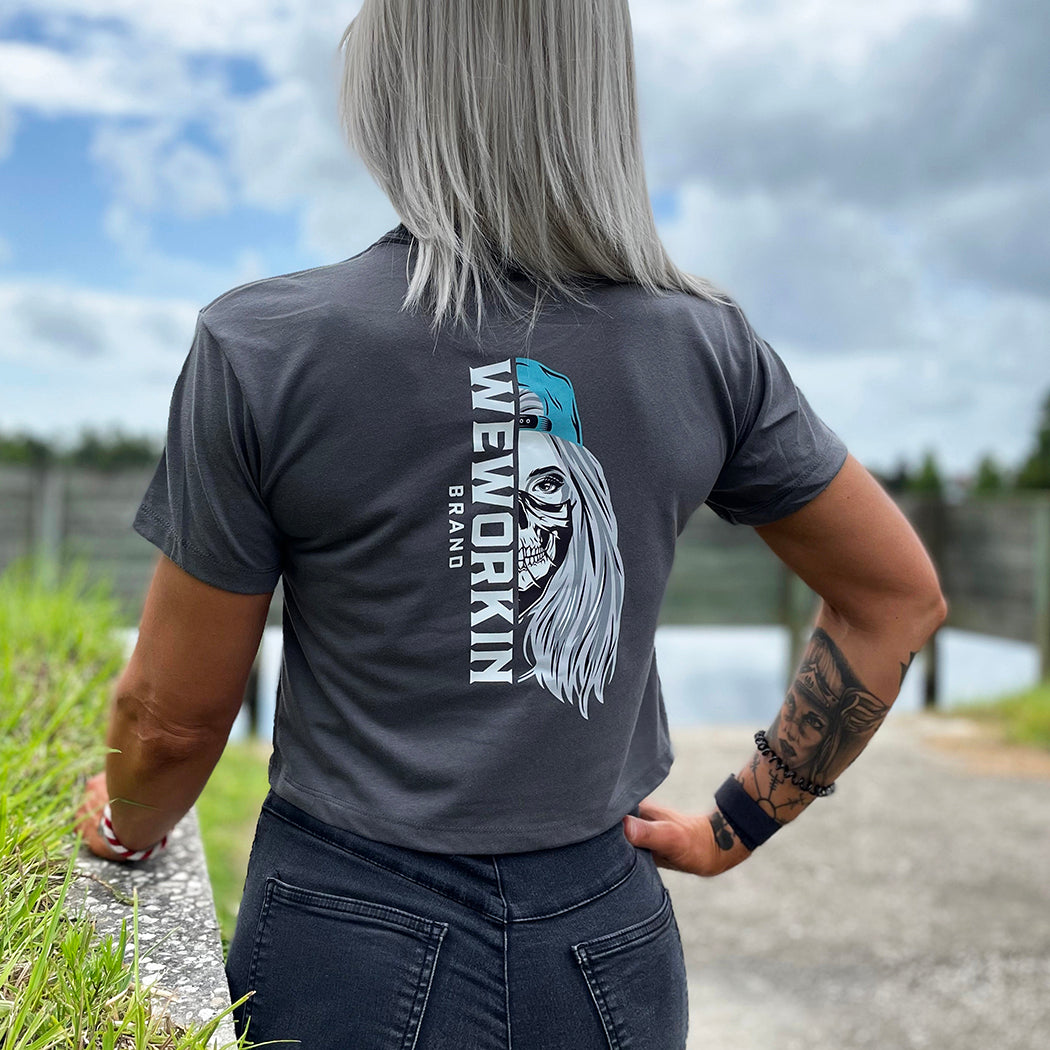 Woman wearing a We Workin Women's short-sleeve cropped tee in dark grey—with a large imprint of our "WEWORKIN BRAND vertical text and Half Skull Woman with Hat" design in Black/White/Grey (hat graphic highlighted in Teal ink.)