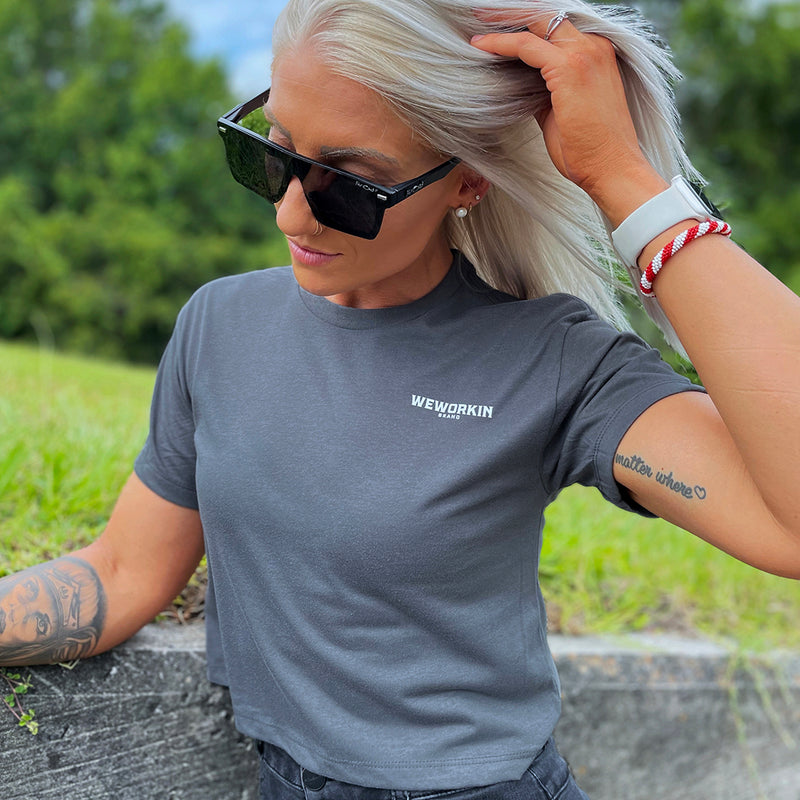 Front of a woman wearing a We Workin Women's short-sleeve cropped tee in white—with a small imprint of our "WEWORKIN BRAND" icon on the upper left chest in White ink.