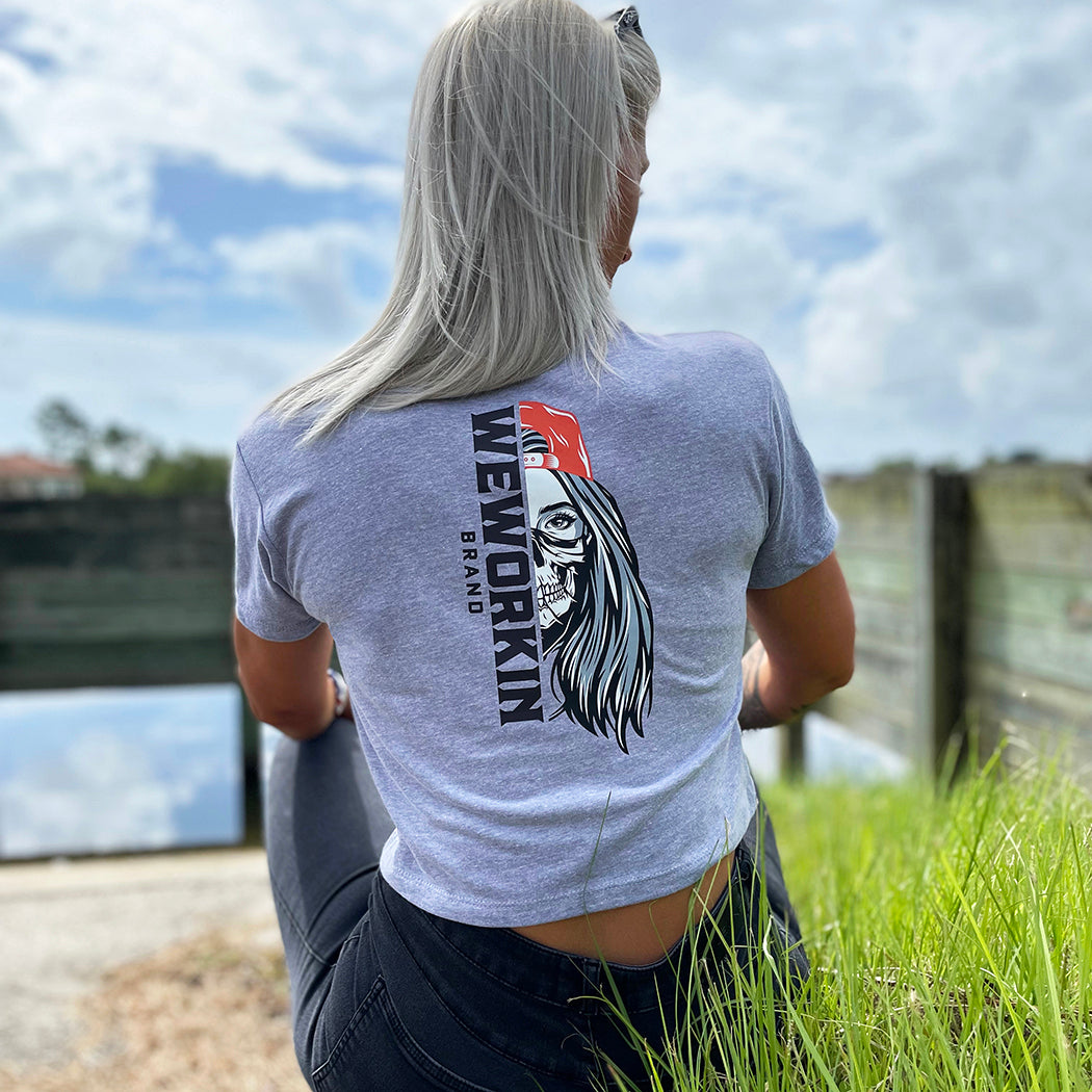 Woman wearing a We Workin Women's short-sleeve cropped tee in heather grey—with a large imprint of our "WEWORKIN BRAND vertical text and Half Skull Woman with Hat" design in Black/White/Grey (hat graphic highlighted in Orange ink.)