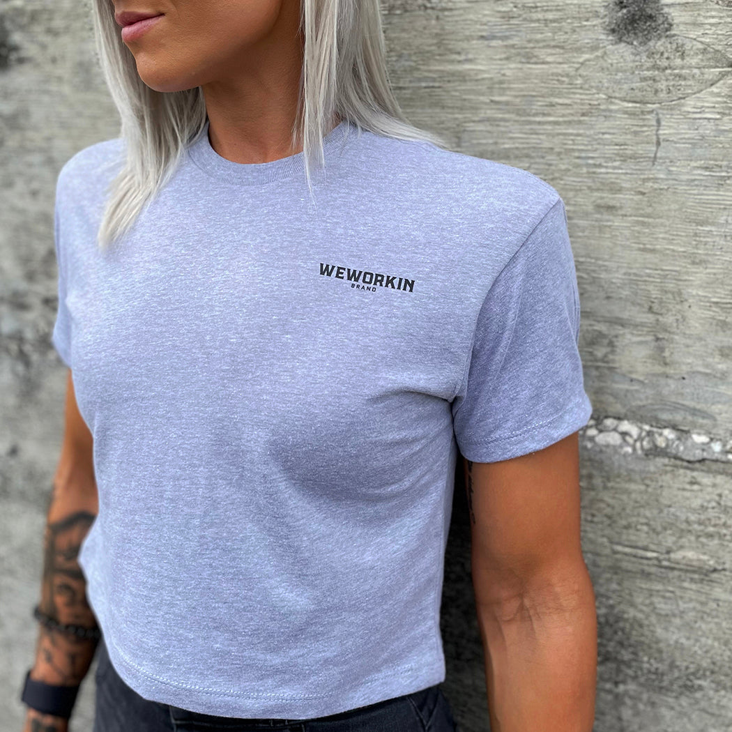 Front of a woman wearing a We Workin Women's short-sleeve cropped tee in heather grey—with a small imprint of our "WEWORKIN BRAND" icon on the upper left chest in Black ink.