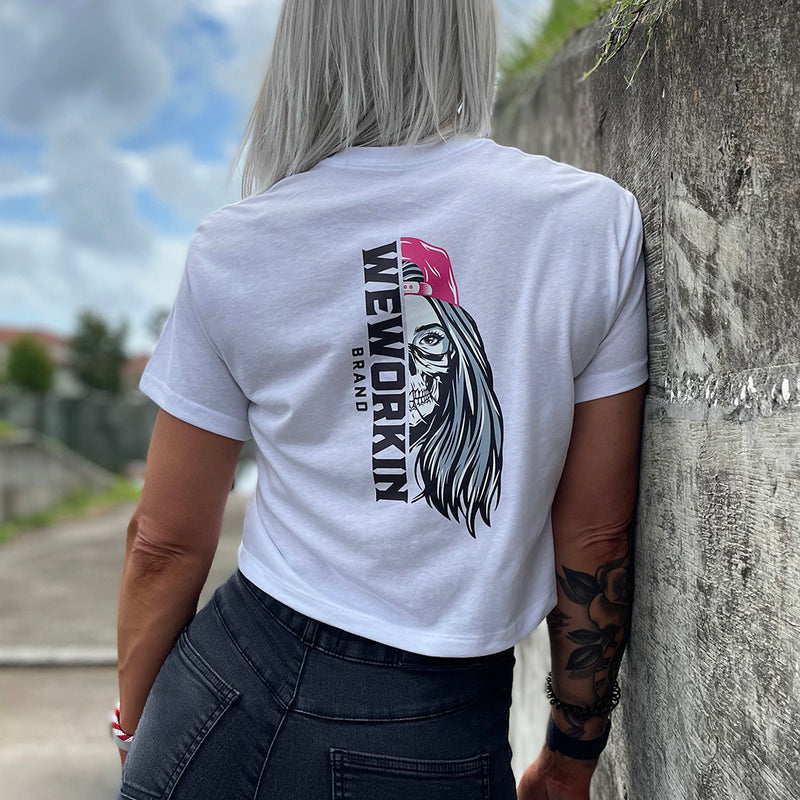 Woman wearing a We Workin Women's short-sleeve cropped tee in white—with a large imprint of our "WEWORKIN BRAND vertical text and Half Skull Woman with Hat" design in Black/White/Grey (hat graphic highlighted in Bright Pink ink.)