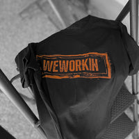 We Workin black graphic tee, boldly screenprinted with our original design "WEWORKIN Dirt Stamp" graphic, on the full front chest, in brown ink laying on a step ladder.