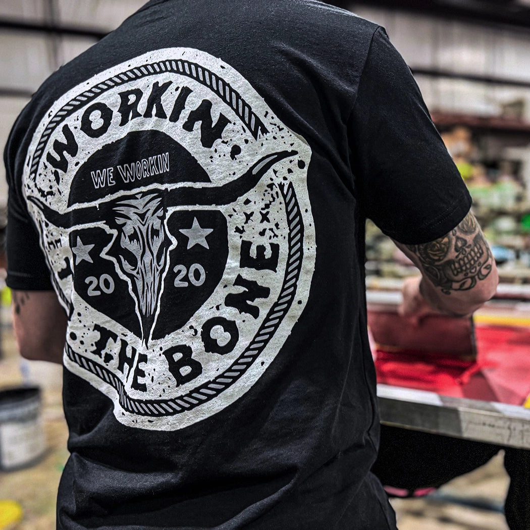 Man pictured from back, wearing a "WORKIN TO THE BONE" WW black tee. "WORKIN TO THE BONE" text circular graphic with a cow skull in the center is printed large in the center/upper back, in grey and white ink.