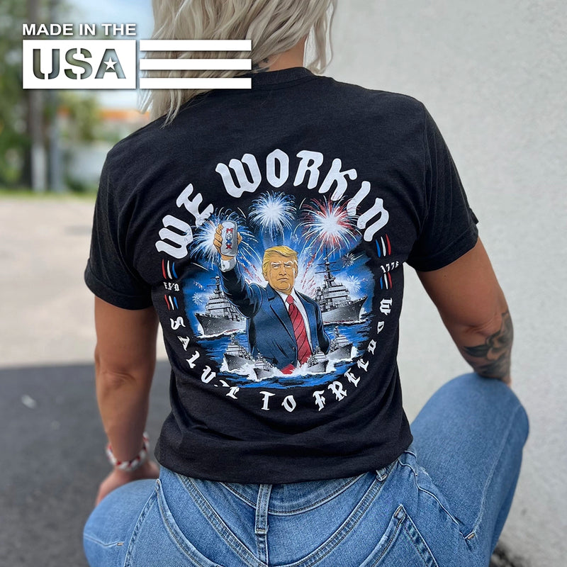 Woman wearing a WW black tee MADE IN THE USA, shown from back. "WE WORKIN—SALUTE TO FREEDOM" text circling around a President Trump/fireworks "freedom concept" image in center. Graphic is printed large on the centerback. Printed in full color/patriotic inks.