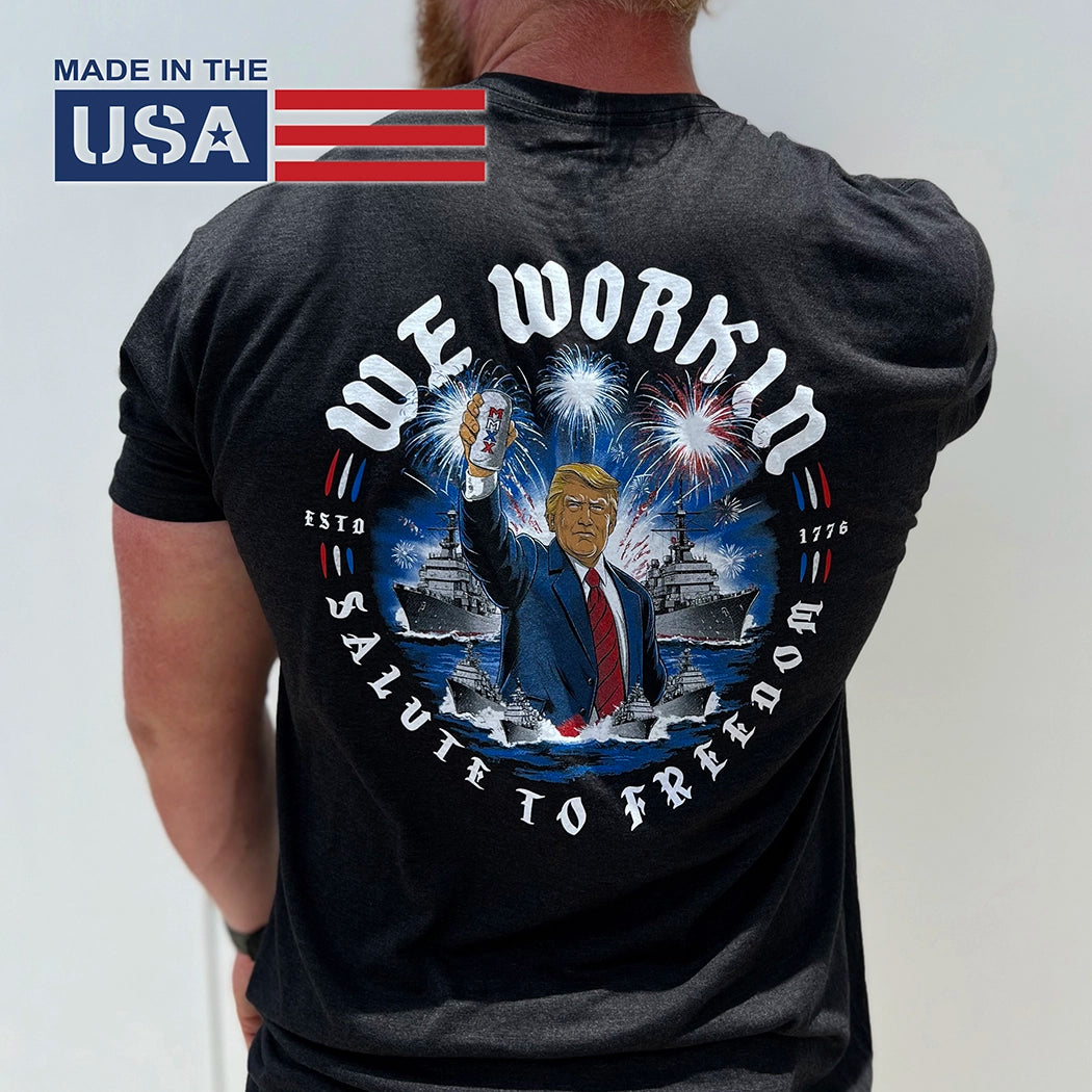 Man wearing a WW black tee MADE IN THE USA, shown from back. "WE WORKIN—SALUTE TO FREEDOM" text circling around a President Trump/fireworks "freedom concept" image in center. Graphic is printed large on the centerback. Printed in full color/patriotic inks.