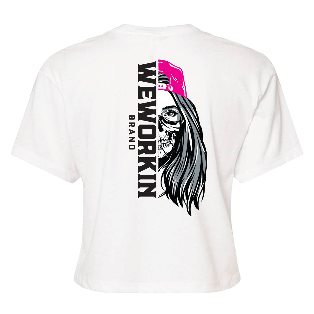 Back of a We Workin Women's short-sleeve cropped tee in white—with a large imprint of our "WEWORKIN BRAND vertical text and Half Skull Woman with Hat" design in Black/White/Grey (hat graphic highlighted in Bright Pink ink.)