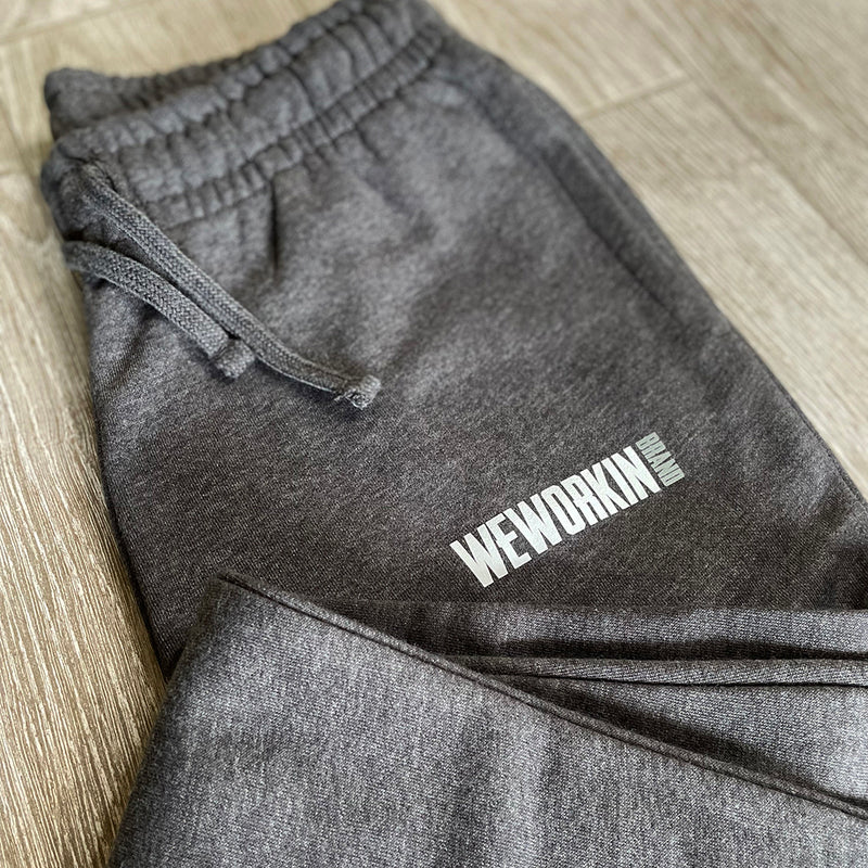 Front view of the We Workin women's joggers in Heathered Charcoal Grey, folded to show the printed logo. "WEWORKIN BRAND" block logo is subtly printed just below the front left pocket area in white and grey ink. [Graphic approx 3.5" wide.]