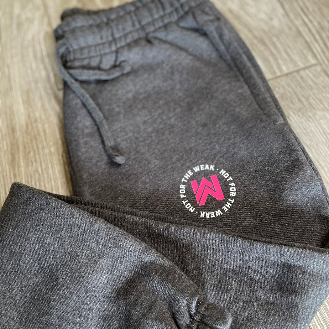 Front view of the We Workin women's joggers in Heathered Charcoal Grey, folded to show the printed logo. "NOT FOR THE WEAK" white text encircling the WW icon logo in bright pink, subtly printed just below the front left pocket area. [Design approx 2.25" wide.]