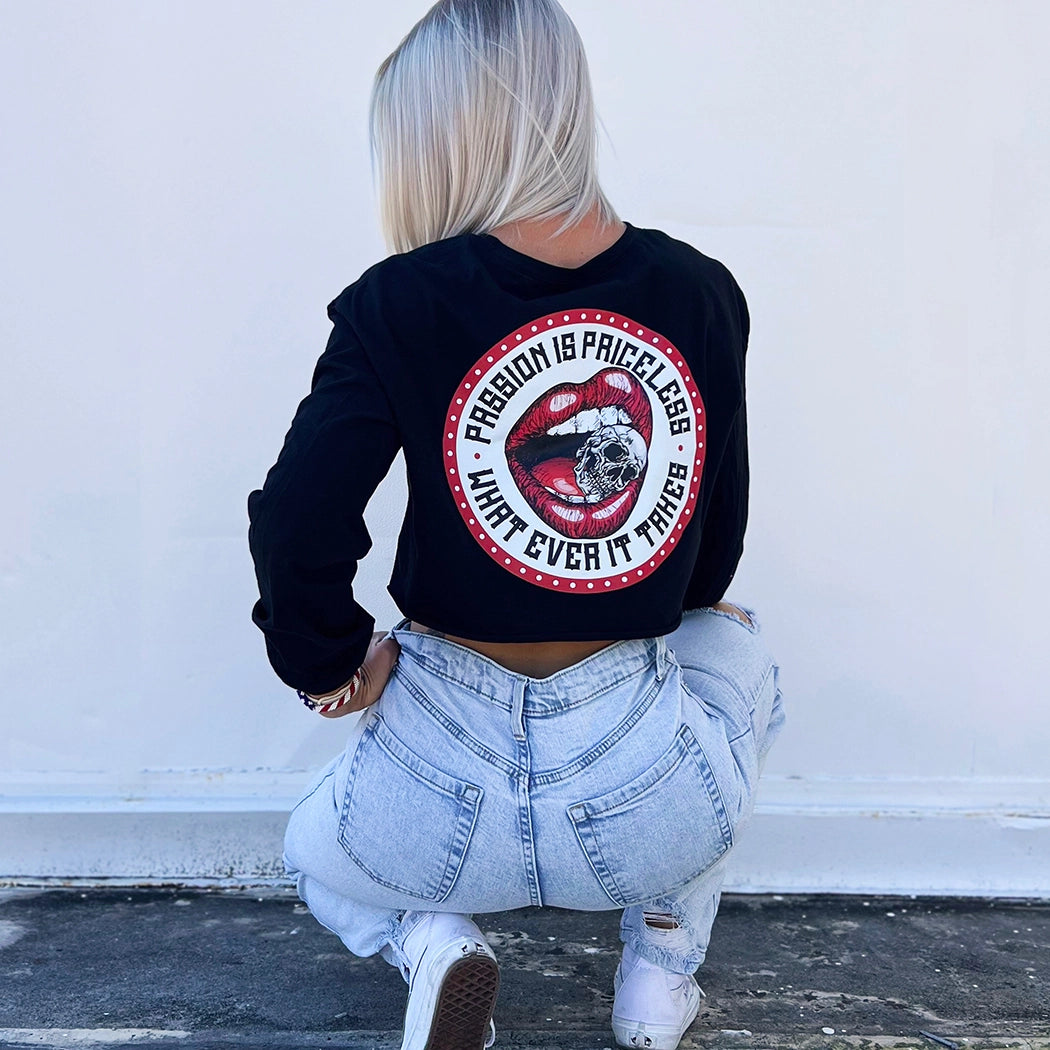 Woman outside, pictured from back, wearing a We Workin women’s cropped long sleeve shirt in black. Full color graphic on back includes a circular red-beaded border on white background with red lips-mouth "crunching down" on a skull between teeth in the center, the text "PASSION IS PRICELESS • WHAT EVER IT TAKES" around graphic in black ink.