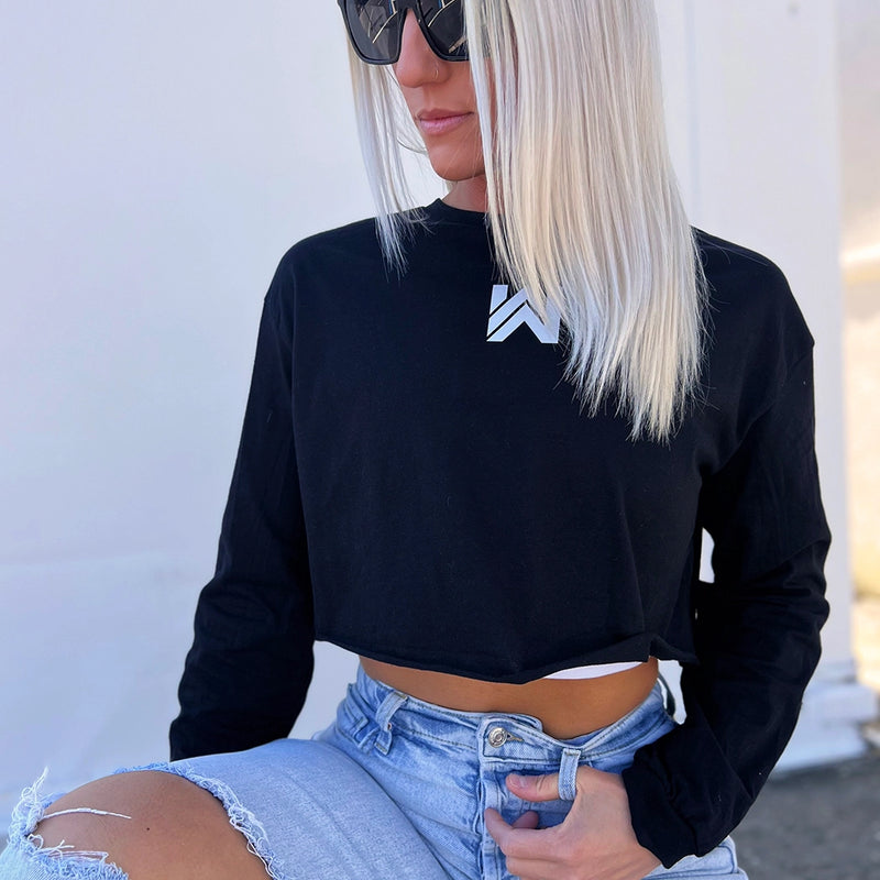 Woman, outside, pictured from front, wearing a We Workin women’s cropped long sleeved tee in black. Small printed WW icon on center chest near neckline, in white ink.