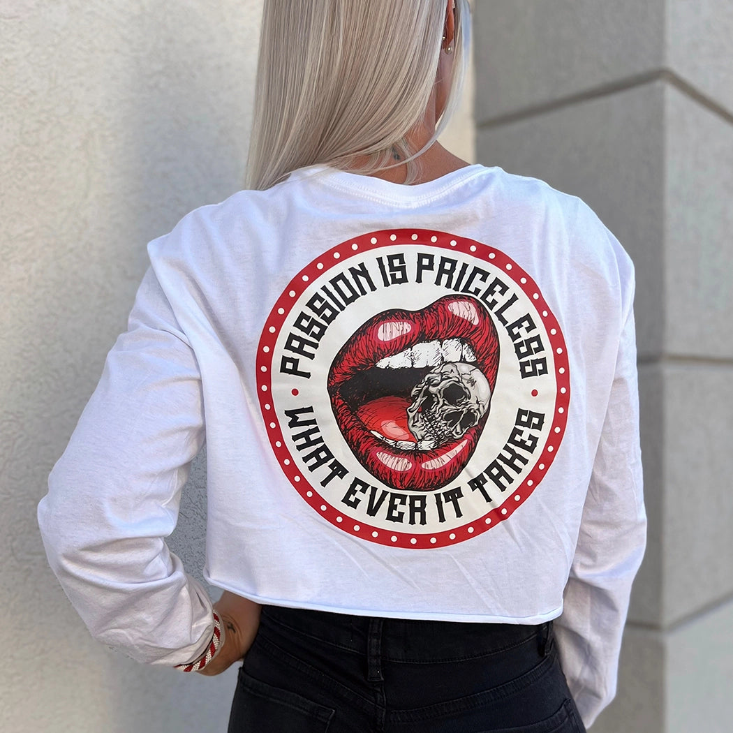 Woman outside, pictured from back, wearing a We Workin women’s cropped long sleeve shirt in white. Full color graphic on back includes a circular red-beaded border on white background with red lips-mouth "crunching down" on a skull between teeth in the center, the text "PASSION IS PRICELESS • WHAT EVER IT TAKES" around graphic in black ink.