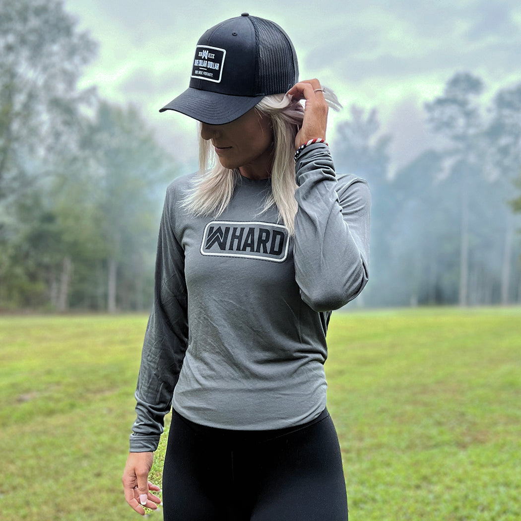 Woman pictured outdoors, wearing a WE WORKIN lightweight long sleeve tee in Smoke Grey color. We Workin long sleeve tee in Smoke Grey color. Front is printed with the We Workin WWHARD patch-style icon printed in Black ink, except the "rope" style outline is in white ink. She's also wearing an all black "Blue Collar Dollar" patch hat in classic-style (Richardson 112).