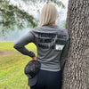 Woman pictured outdoors from the back, wearing a WE WORKIN lightweight long sleeve tee in Smoke Grey color. The back is printed with the tagline "Everybody Wants the Money, Nobody Wants the WORK." in black ink, except the word WORK is in white ink.