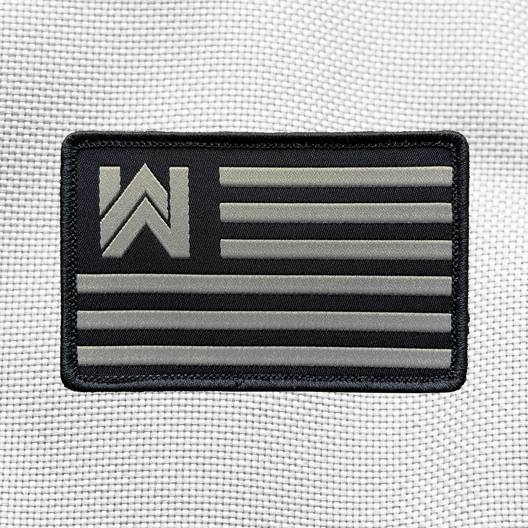 WEWORKIN Branded FLAG on a velcro-backed patch (both the hook and loop sides provided). [1] thread color for the WW and stripes (grey) on a black woven background, with black merrowed border. 3.5" wide Woven patch displayed on a light grey canvas background.