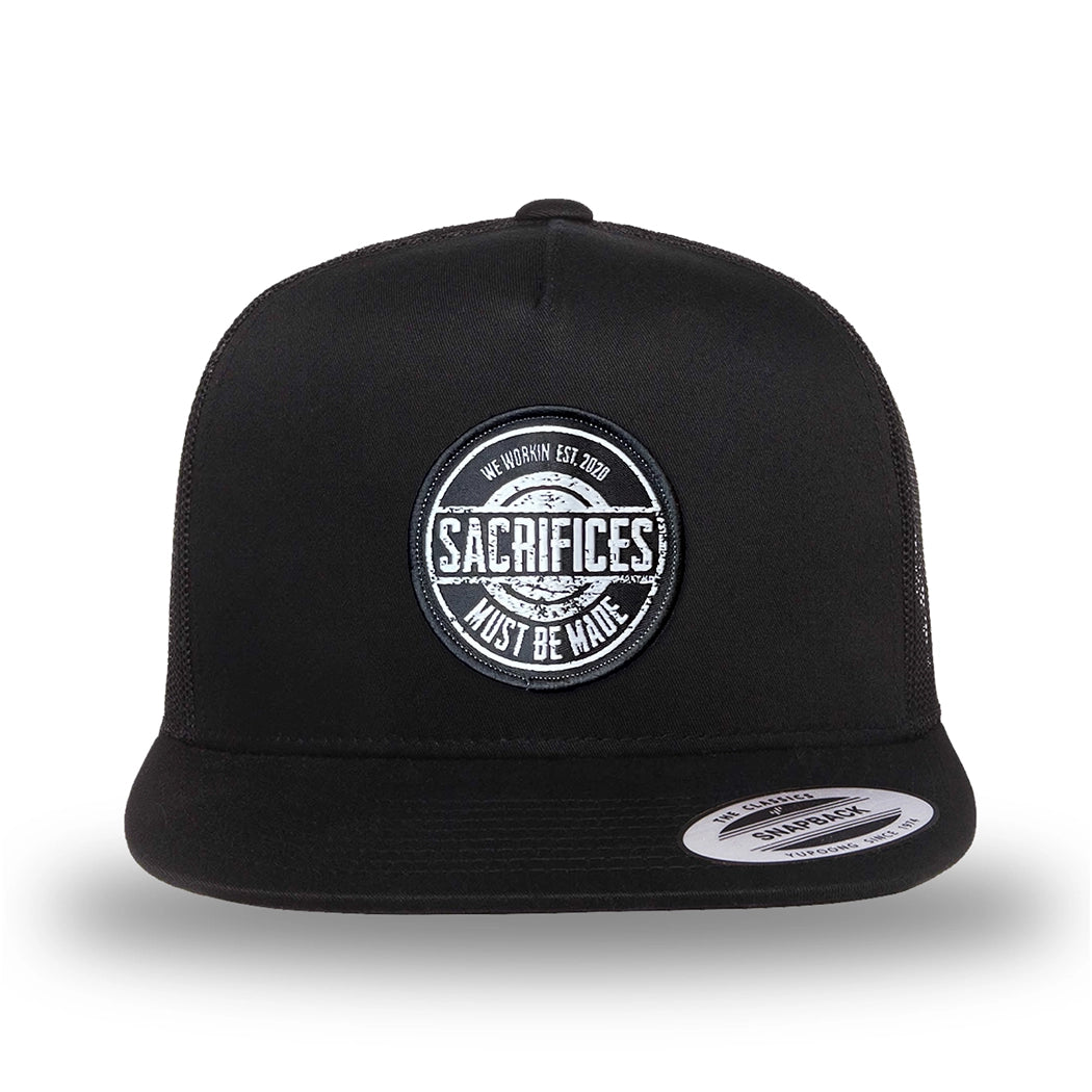 All black, high-profile, WeWorkin hat—snapback, 5-panel classic trucker, mesh sides/back style. WeWorkin "SACRIFICES MUST BE MADE" circular woven patch is centered on the front panel.
