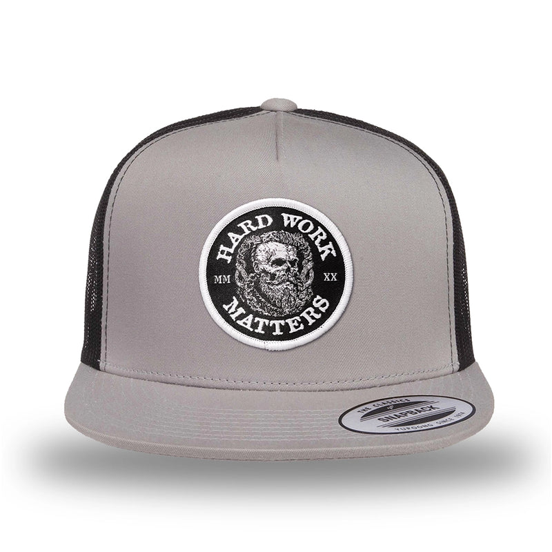 Silver/Black two-tone WeWorkin hat—snapback, 5-panel classic trucker, mesh sides/back style. HARD WORK MATTERS woven patch with white merrowed edge, on a black background with HARD WORK MATTERS text encircling a Viking-style skull center graphic with MM XX on the left and right respectively—patch is centered on the front panels.