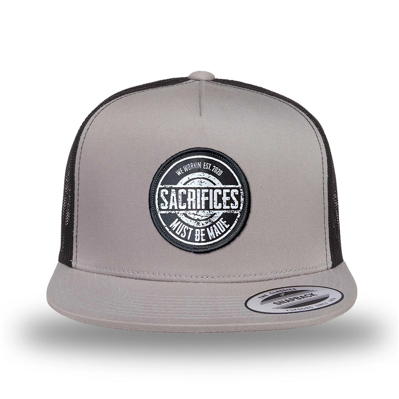 Silver/Black two-tone WeWorkin hat—snapback, 5-panel classic trucker, mesh sides/back style. WeWorkin "SACRIFICES MUST BE MADE" circular woven patch is centered on the front panel.
