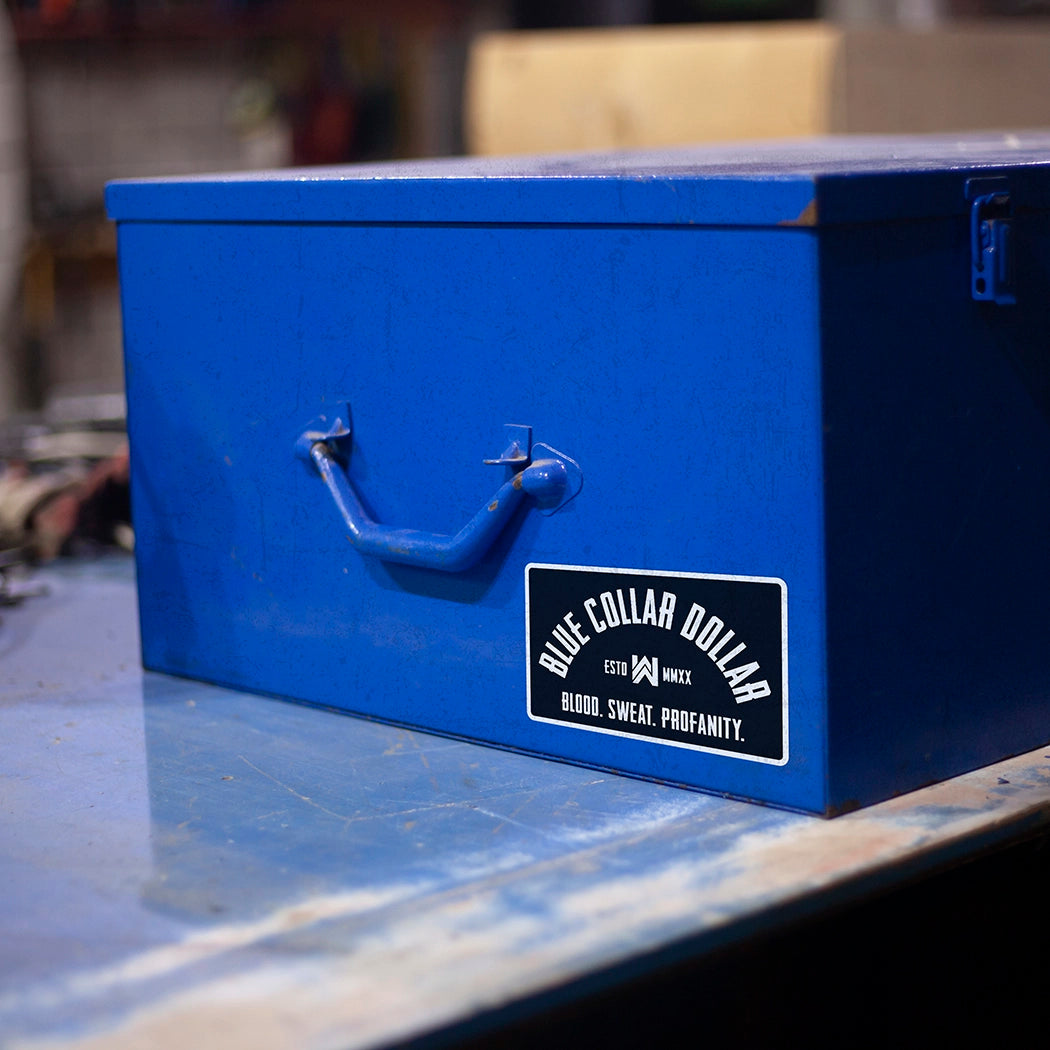 "BLUE COLLAR DOLLAR. Blood. Sweat. Profanity." arch design die cut sticker on a toolbox on top of a work bench.