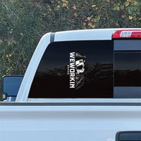 Cropped in image of a LARGE size WEWORKIN BRAND Black/White Skull decal 11"H—Custom die-cut Direct Transfer window sticker, on rear window (Left side, centered) of a Chevy truck. Foliage in background.