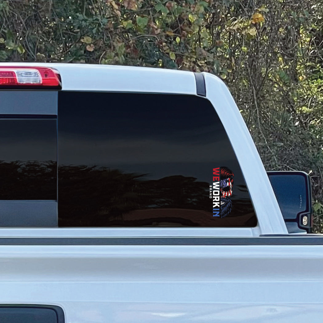 Cropped in image of a SMALL size WEWORKIN BRAND Patriotic Skull 6.75"H—Custom die-cut Direct Transfer window sticker in multi-color, on rear window (Right side, lower right corner) of a Chevy truck. Foliage in background.