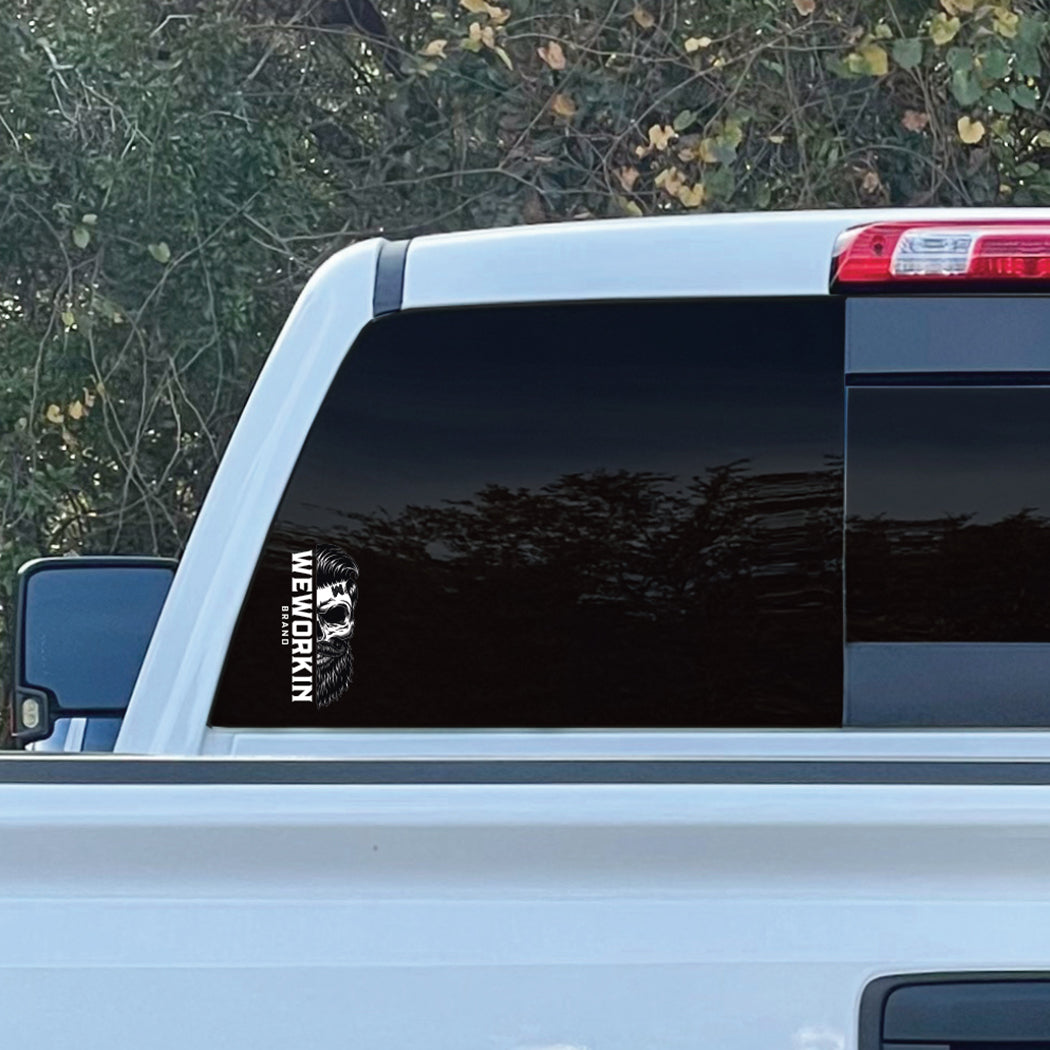 Cropped in image of a SMALL size WEWORKIN BRAND Black/White Skull decal 6.75"H—Custom die-cut Direct Transfer window sticker, on rear window (Left side, lower left corner) of a Chevy truck. Foliage in background.