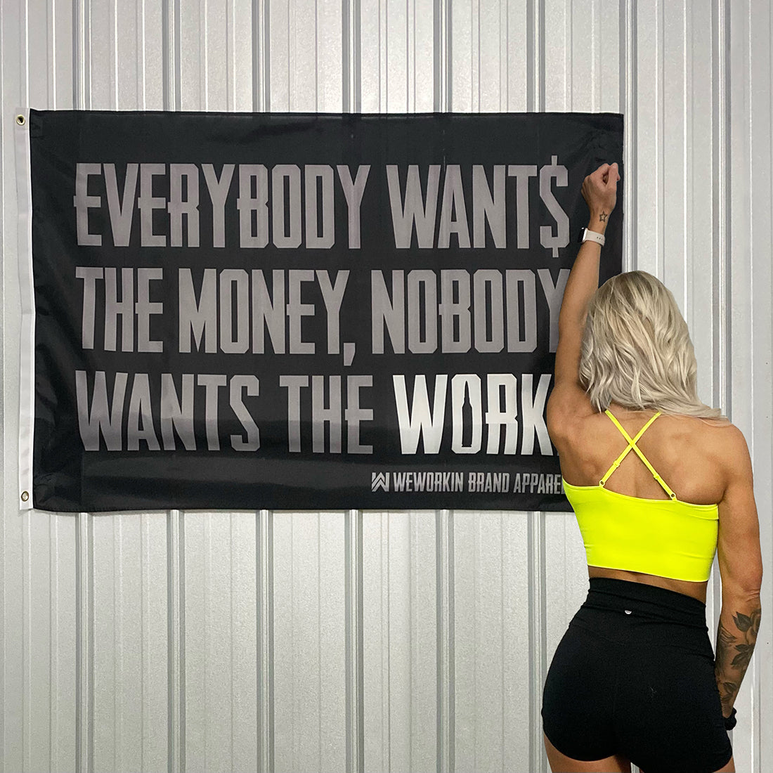 WeWorkin Brand "Everybody Wants the Money, Nobody Wants the WORK." flag displayed on a corrugated metal wall. Each flag measures 5'w x 3'h, black background with all grey letters except for the word "WORK" which is in white. White, double-stitched, thicker left edge for durability, (2) gold grommets (one at top left and one at bottom left corners).