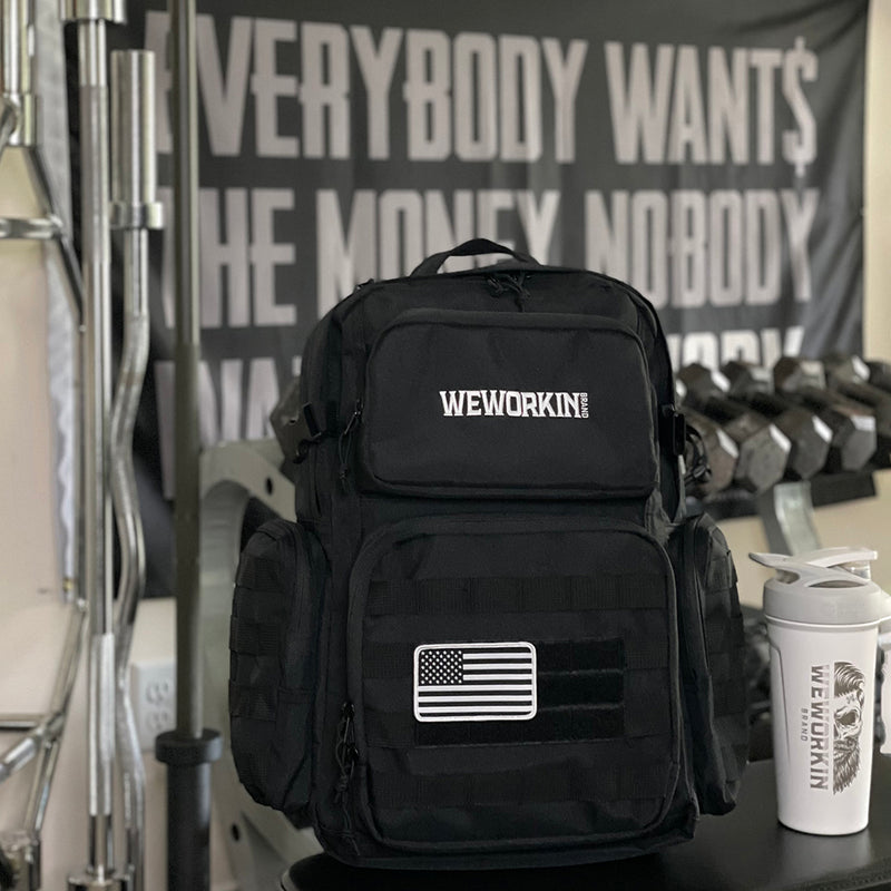 Stealth Black tactical backpack pictured from front in a gym. WEWORKIN BRAND logo embroidered in white thread on the top front pocket center. Two side zippered accessory pockets with daisy chain, Top front zippered pocket embroidered, Web carry handle, Lower front zippered pocket with daisy chain and loop panel for badges and patches, American flag patch shown on bag (sold separately).