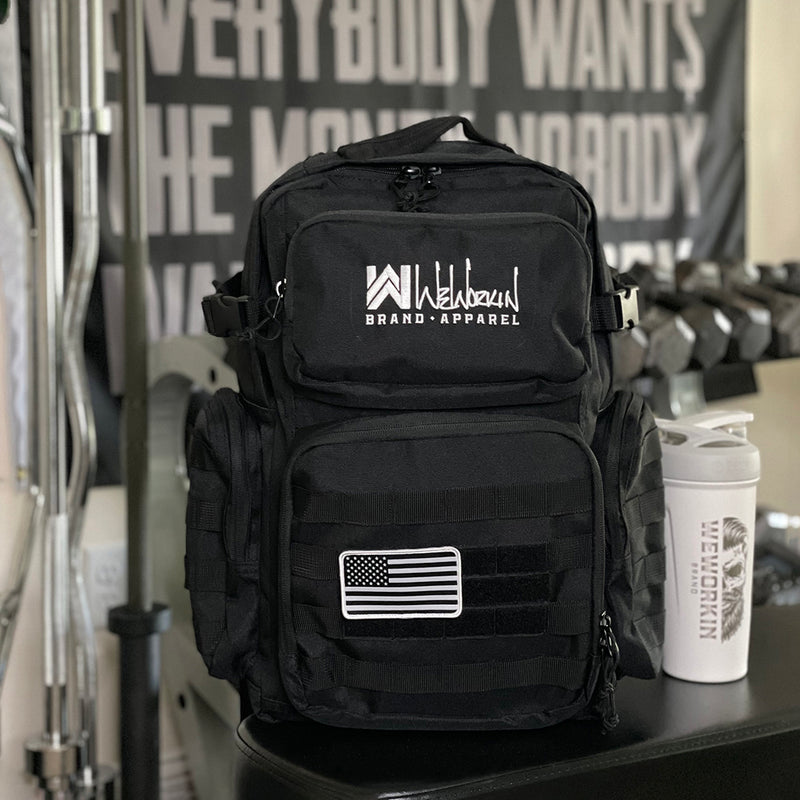 Stealth Black tactical backpack pictured from front in a gym. WW Script logo embroidered in white thread on the top front pocket center. Two side zippered accessory pockets with daisy chain, Top front zippered pocket embroidered, Web carry handle, Lower front zippered pocket with daisy chain and loop panel for badges and patches, American flag patch shown on bag (sold separately).