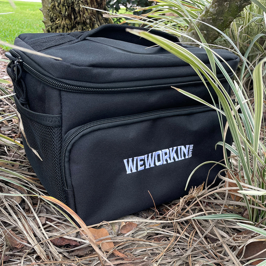 Stealth Black insulated cooler bag pictured from front on ground outside. WEWORKIN BRAND logo embroidered in white thread on the front zippered pocket. 600D polyester canvas, Water-resistant PEVA lining (Heat-sealed), Web carry handle on top, Removable/adjustable shoulder strap, Mesh pocket on side.