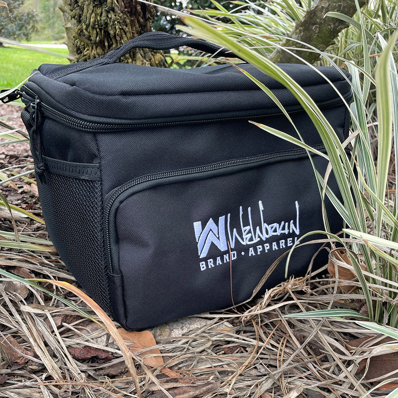 Stealth Black insulated cooler bag pictured from front on ground outside. WW Script logo embroidered in white thread on the front zippered pocket. 600D polyester canvas, Water-resistant PEVA lining (Heat-sealed), Web carry handle on top, Removable/adjustable shoulder strap, Mesh pocket on side.