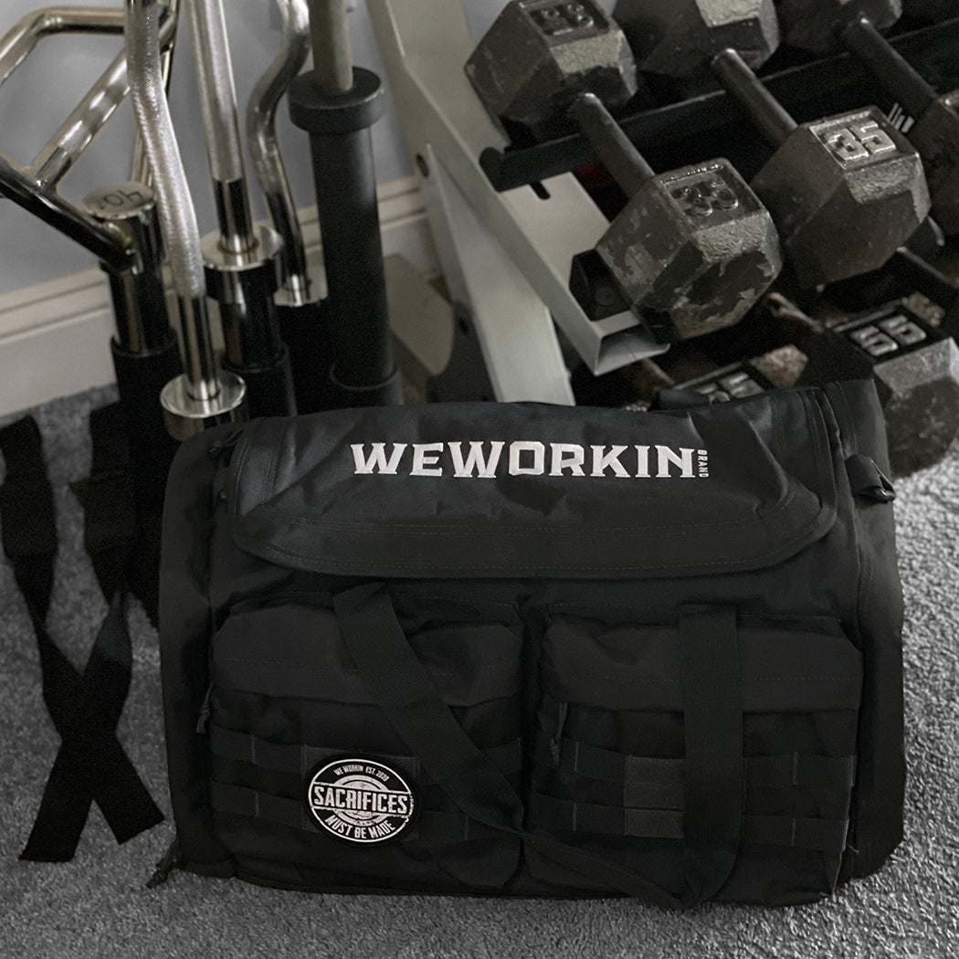 Stealth Black tactical duffle bag pictured from front on gym floor, beside a weight rack. WEWORKIN BRAND logo embroidered in white thread on the top panel. 600D polyester canvas, Covered zipper D-shaped main compartment, [2] zippered end pockets, [2] Front zippered pockets with daisy chain/loop panels for patches (WW SACRIFICES patch shown on duffle bag, all patches sold separately).