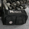 Stealth Black tactical duffle bag pictured from front on gym floor, beside a weight rack. WW Script logo embroidered in white thread on the top panel. 600D polyester canvas, Covered zipper D-shaped main compartment, [2] zippered end pockets, [2] Front zippered pockets with daisy chain/loop panels for patches (WW SACRIFICES patch shown on duffle bag, all patches sold separately).