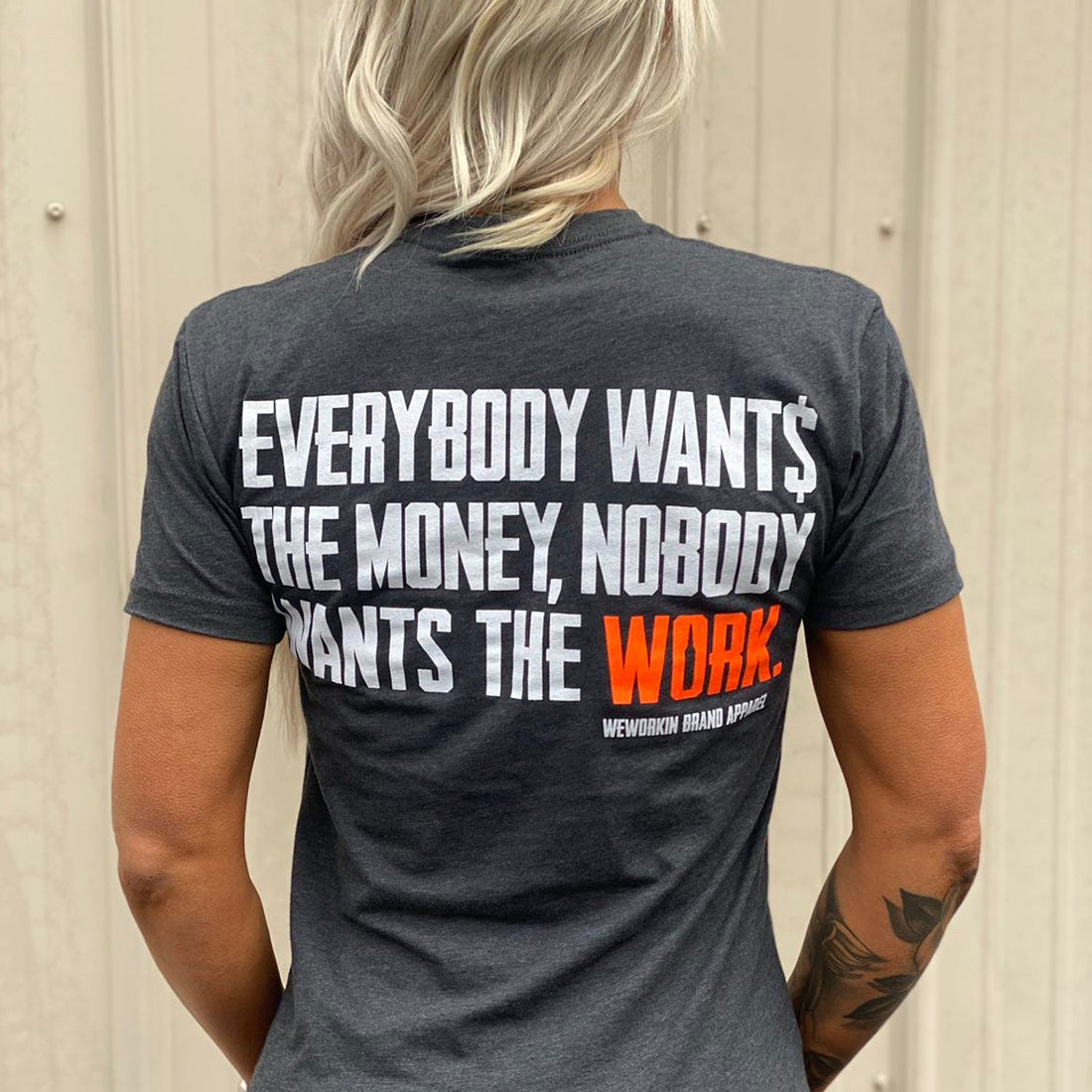 Woman pictured from back wearing a WW Charcoal Heather Grey Graphic Tee. "EVERYBODY WANT$ THE MONEY, NOBODY WANTS THE WORK" printed in bold on full back width in white ink, except the word WORK is printed in Neon Orange for emphasis. (WEWORKIN BRAND APPAREL printed small and lower right just below the full back imprint.)