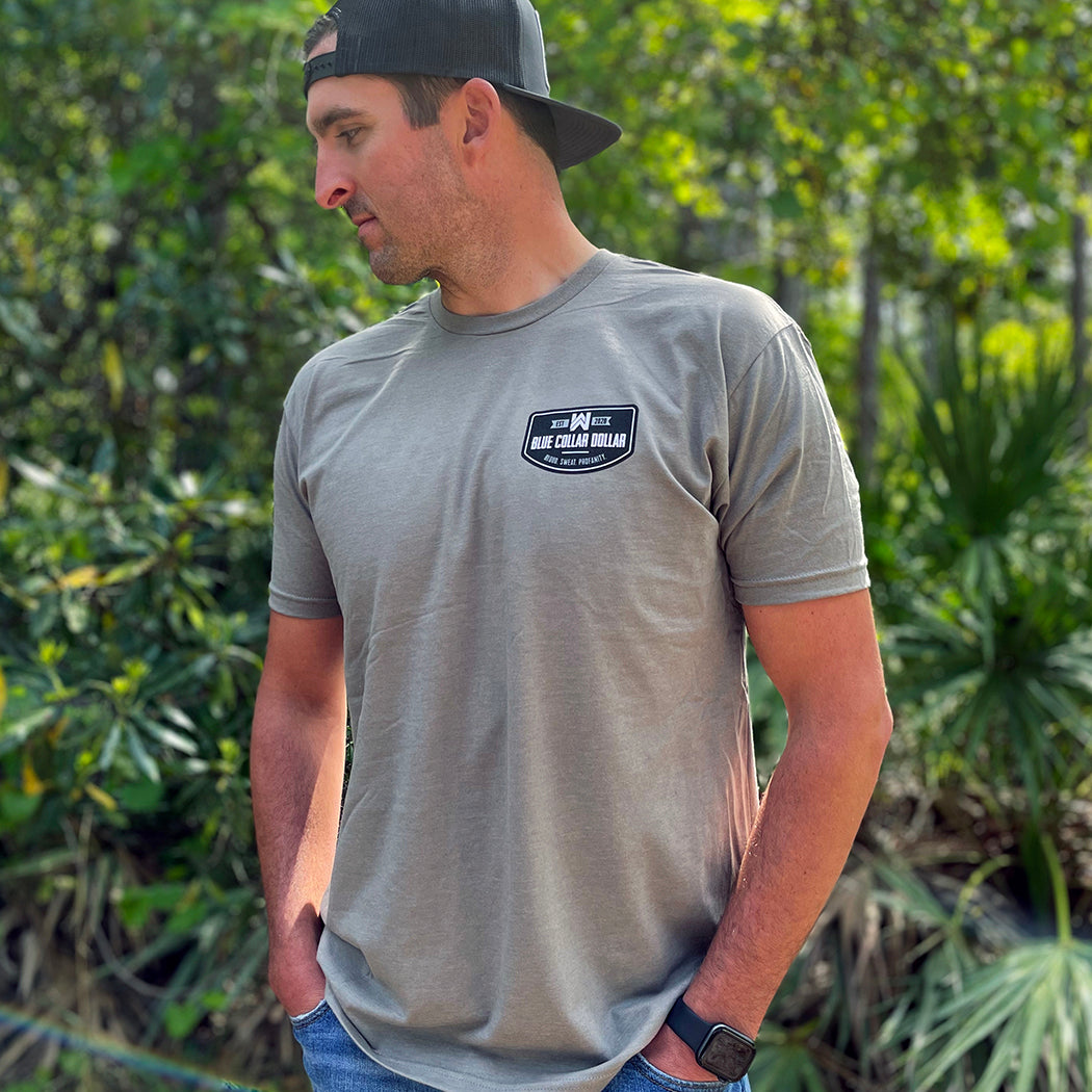 Man, from front, wearing a We Workin graphic tee in Stone Grey color. Short sleeve shirt printed with We Workin designed emblem graphic—"BLUE COLLAR DOLLAR. BLOOD. SWEAT. PROFANITY." printed on the left chest "pocket area" (in black, white and grey inks). Man is also wearing a WW Charcoal and Black Flatbill hat with WW large patch on the front panel.