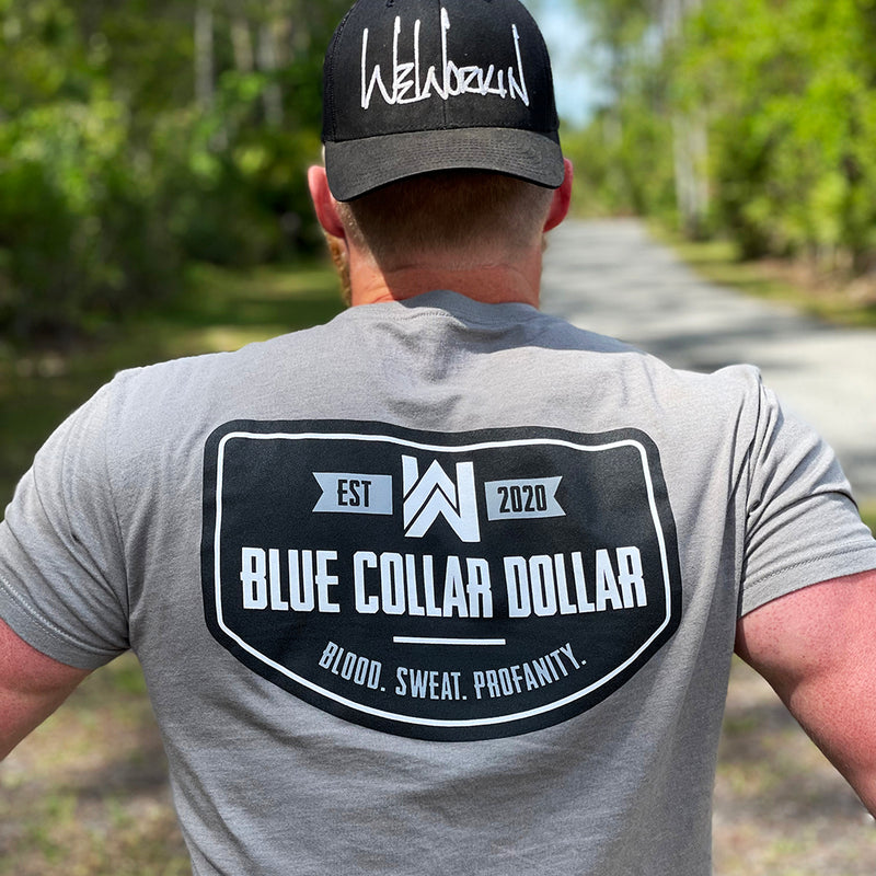 Man, from back, wearing a We Workin graphic tee in Stone Grey color. Short sleeve shirt printed with We Workin designed emblem graphic—"BLUE COLLAR DOLLAR. BLOOD. SWEAT. PROFANITY." printed boldly across the full back (in black, white and grey inks). Man is also wearing a stealth black WW Classic Retro Trucker snapback with the WE WORKIN script logo embroidered in white thread on front panels.