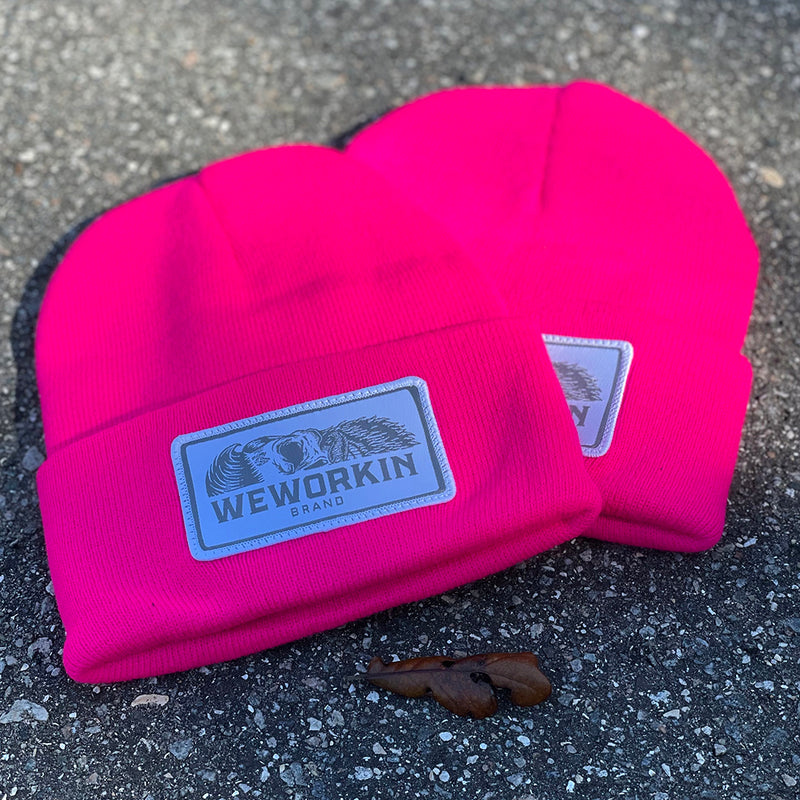 We Workin Neon Pink cuffed beanies laying on asphalt background. Custom 3.5" x 2" patches are sewn on the hats, with the WEWORKIN BRAND text and horizontal half-skull for the design on the patches—woven white background with grey thread for the design and white merrowed-edge border.