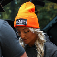 Woman pictured from front wearing a We Workin Brand cuffed beanie in Neon Safety Orange color. Custom 3.5" x 2" patches are sewn on the hats, with the WEWORKIN BRAND text and horizontal half-skull for the design on the patches—woven black background with white thread for the design and black merrowed-edge border.